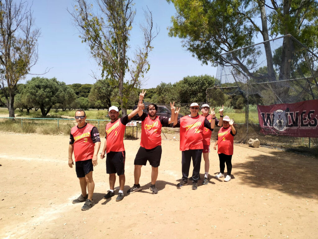 Sis persones equipades per jugar a softbol, amb samarreta taronja i pantalons negres, en un camp de sorra amb alguns arbres al fons, saludant a càmera.