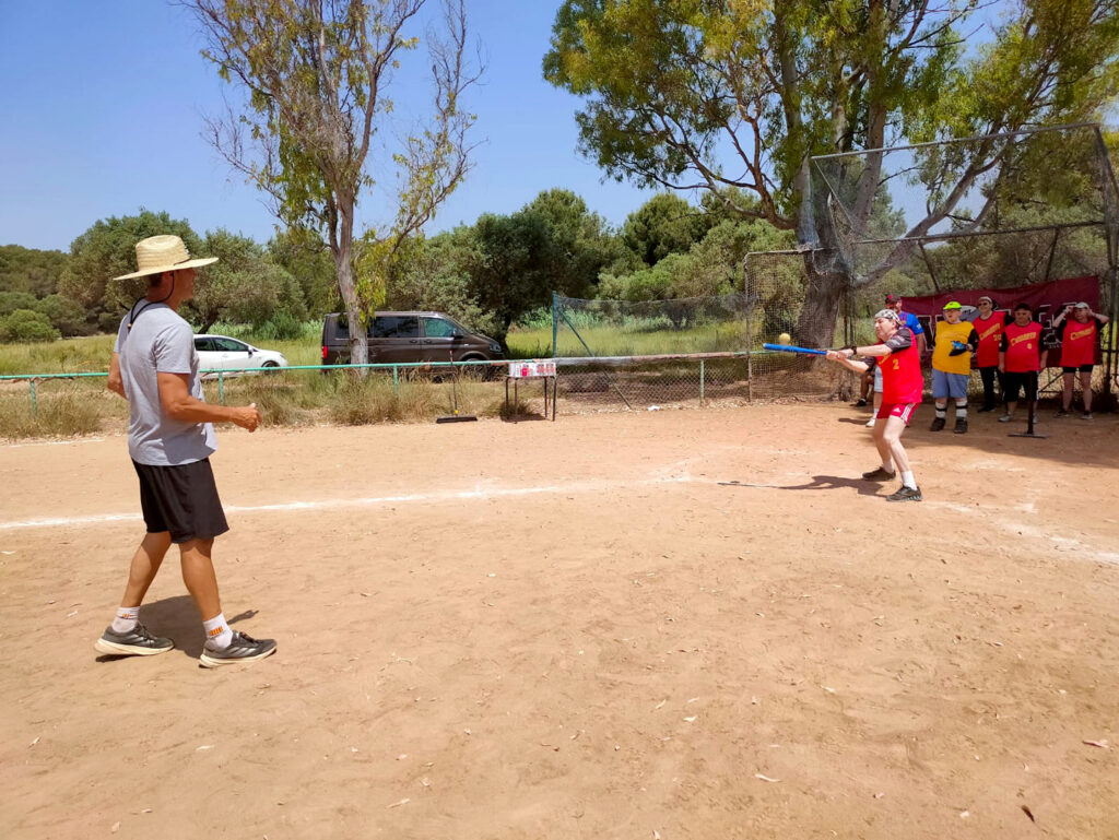 Dues persones jugant a softbol en un camp de sorra, una amb la samarreta taronja i els pantalons negres. La resta de participants son al darrere a l'ombra.
