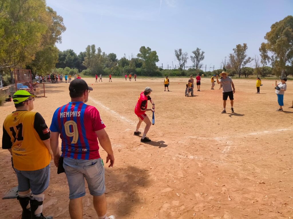 Una desena de persones, d'esquena i cadascuna a una distància diferent de la càmera, jugant un partit de softbol en un camp de sorra.