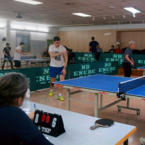 Diferents persones jugant a ping-pong. Destaca una taula de la qual veiem la meitat. A l'extrem inferior esquerre es veu una persona asseguda d'esquenes amb un marcador a sobre de la seva taula.