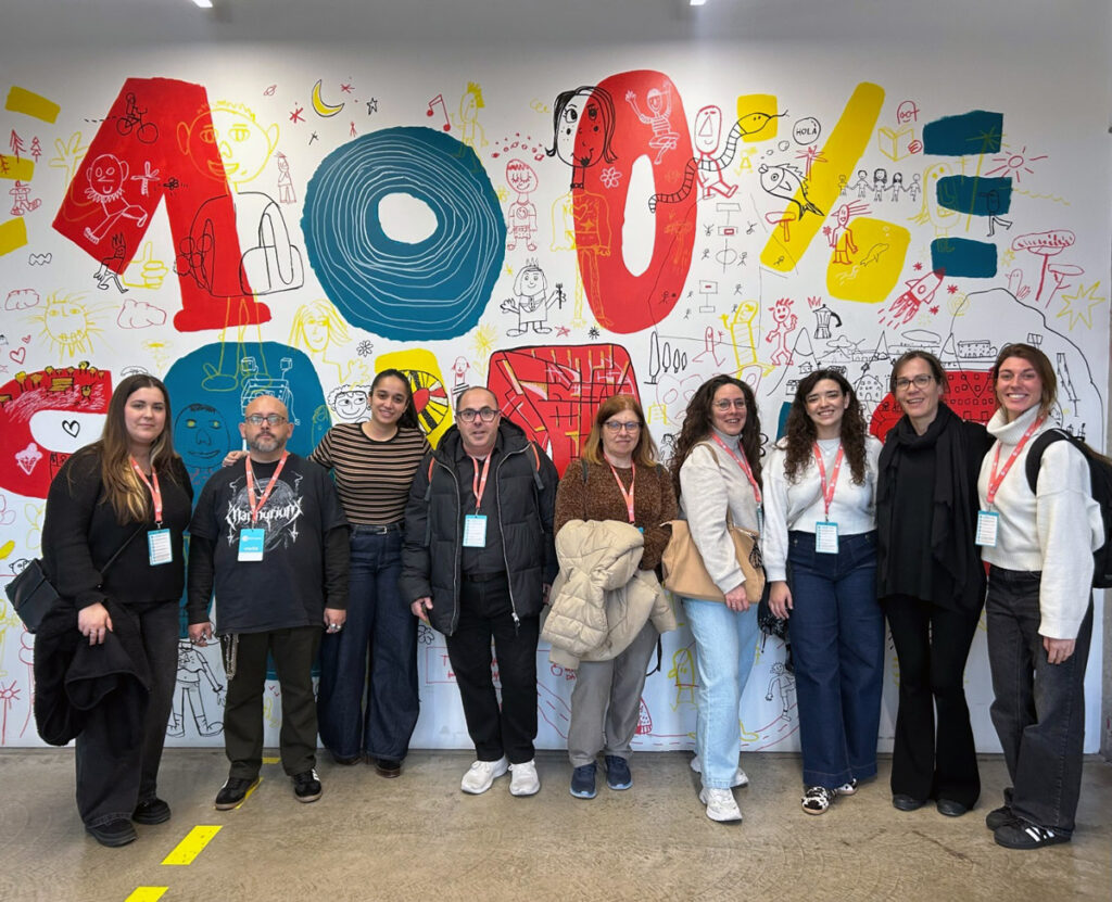 Una foto de grup de nou persones, davant d'una paret pintada amb dibuixos de colors vermell, blau i groc sobre blanc. Es veu el missatge "100%".