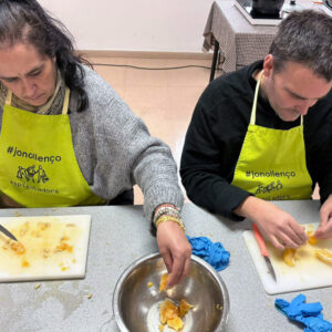 Dues persones amb davantals grocs tallant taronges en una taula i col·locant els trossos en un bol gran. Els davantals són grocs i porten la inscripció "#jonollenço".