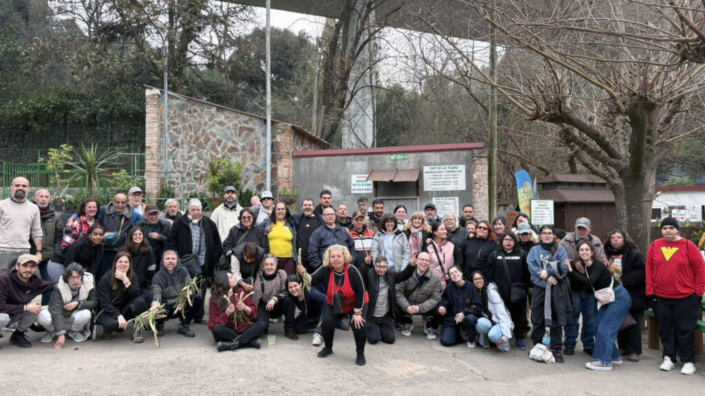 Foto de grup d'unes trenta persones d'edats diferents, en actitud divertida, distribuïdes en tres fileres horitzontals. Al fons hi ha casetes de maons i alguns arbres.