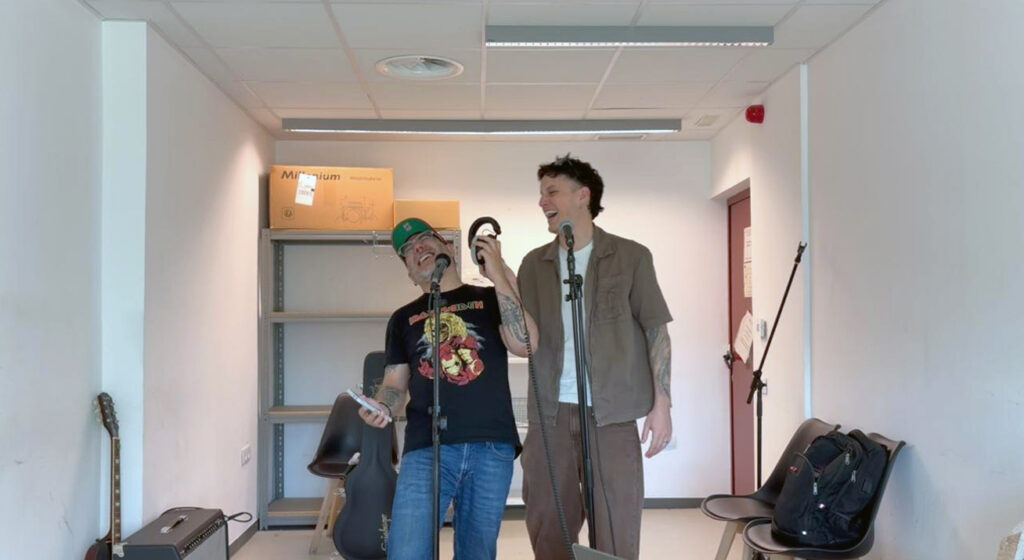 Dues persones de peu rient, en una sala amb ha instruments musicals.