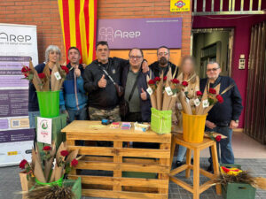 Set persones de diferents edats davant d'una paret de maons amb una senyera vertical i un rètol amb el logo d'Arep. Davant hi ha un palet de fusta i una cadira formant una paradeta amb diverses cistelles de roses de Sant Jordi.