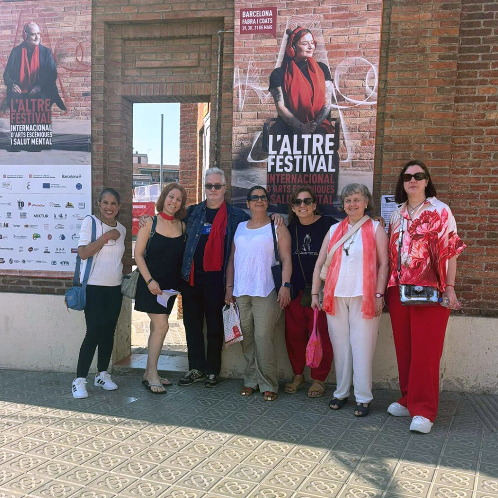 Set persones, cinc de les quals porten roba o accessoris de color vermell, posen per una foto de grup davant d'una paret de maó amb cartelleria de l'Altre Festival.