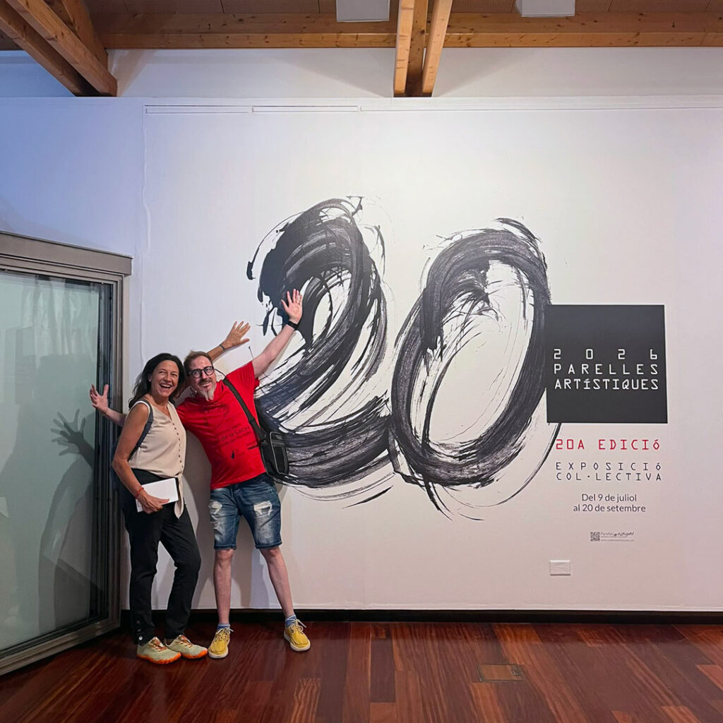 Una dona i un home en una sala d'exposicions, fent-se a un costat per mostrar un número 20 pintat en negre a la paret i uns cartells explicatius.