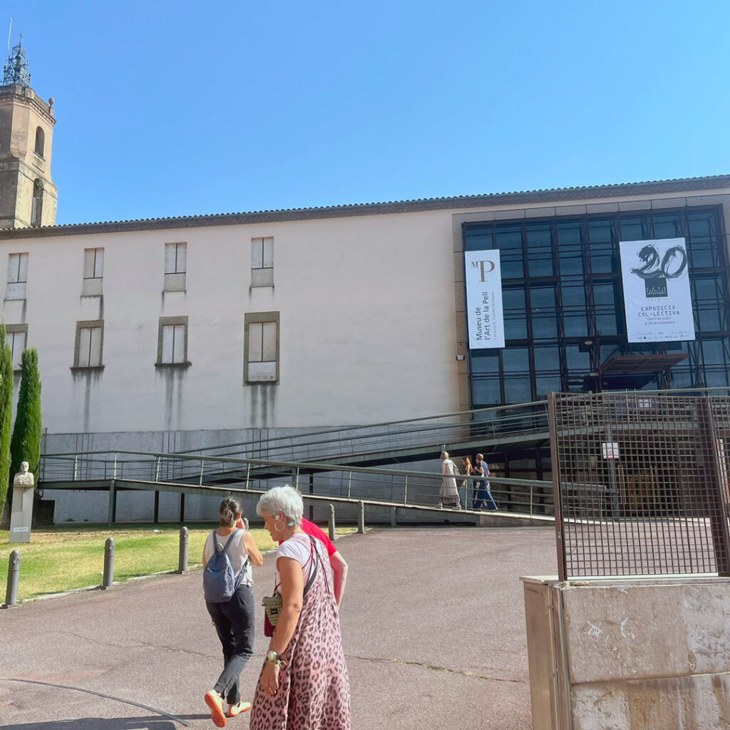 Un parell de persones d'esquena caminen en direcció a un edifici públic d'una sola planta amb la façana blanca i el cel blau de fons.